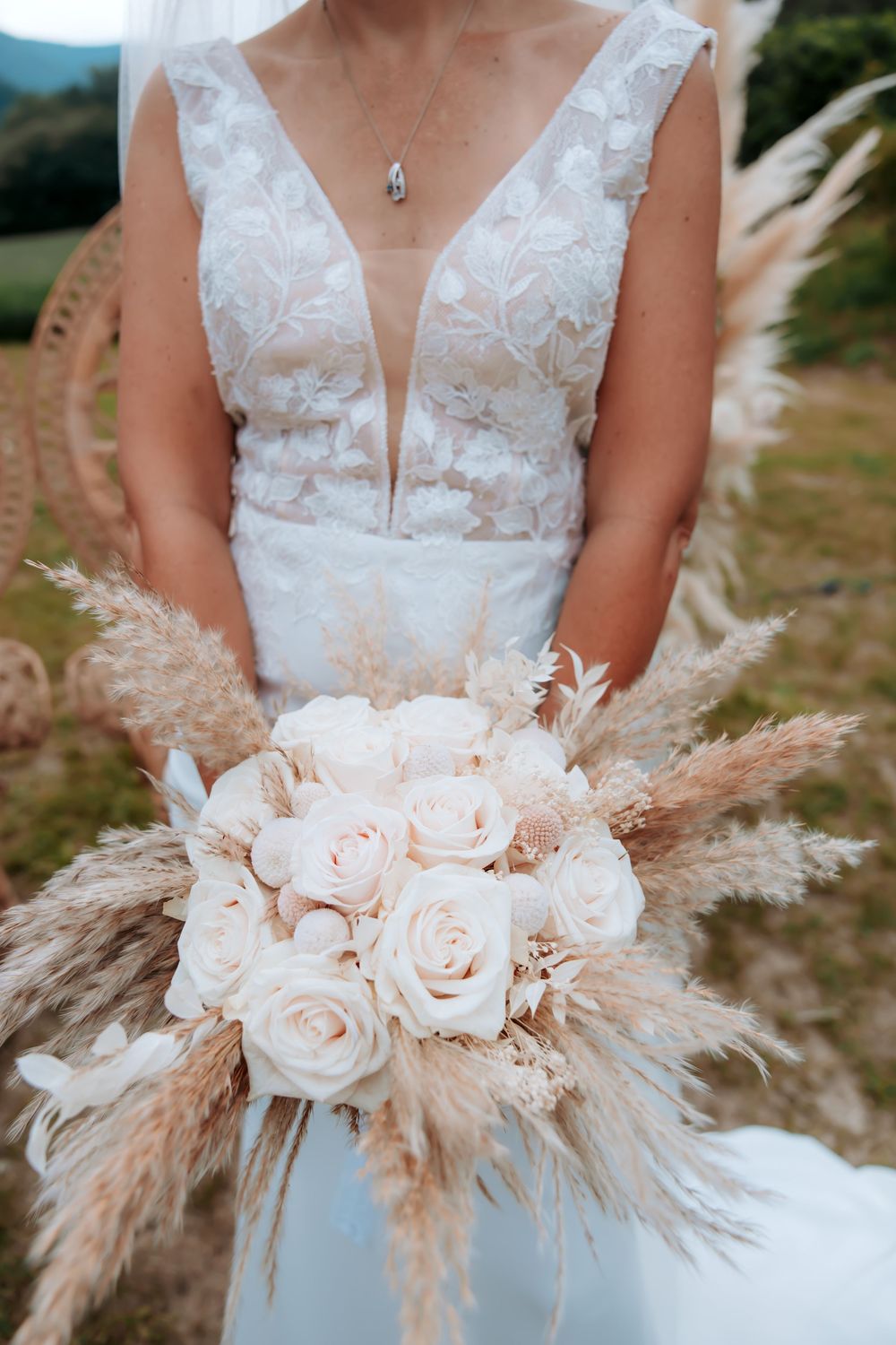 Bride in lace wedding dress holding boho bouquet of white roses and pampas grass outdoors.