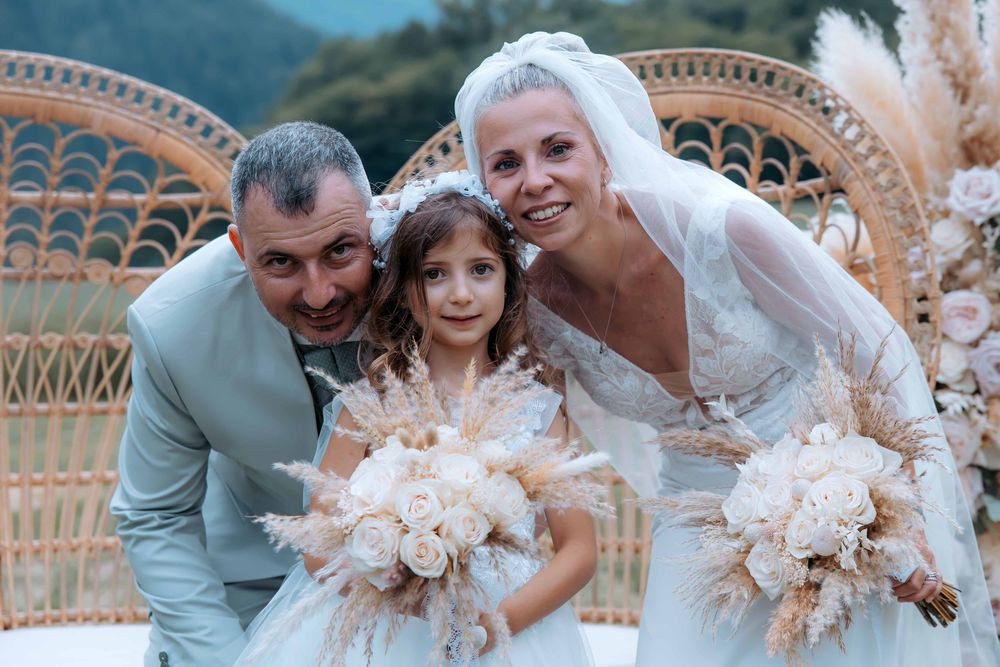 Happy wedding couple with flower girl posing outdoors, bride in white gown holding bouquet, groom in light suit.