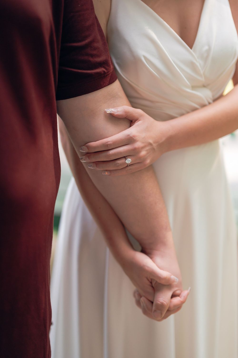 Bride in white gown wearing diamond engagement ring holds hands with partner in burgundy dress.