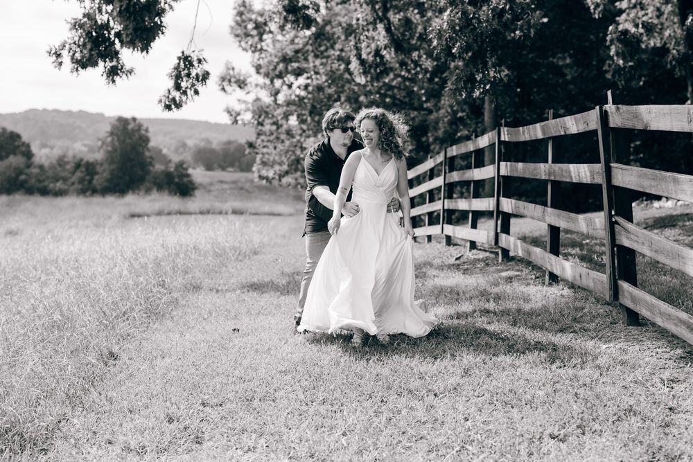 Black and white photo of a bride and groom walking along a wooden fence in a lush countryside field.