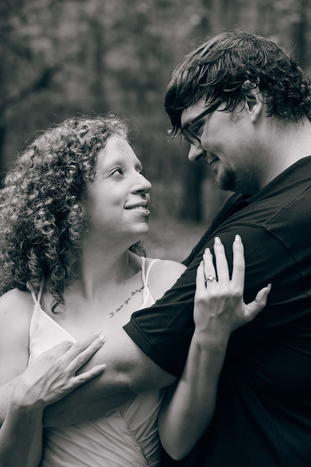 Black and white photo of a smiling couple gazing at each other, woman wearing an engagement ring in a forest setting.