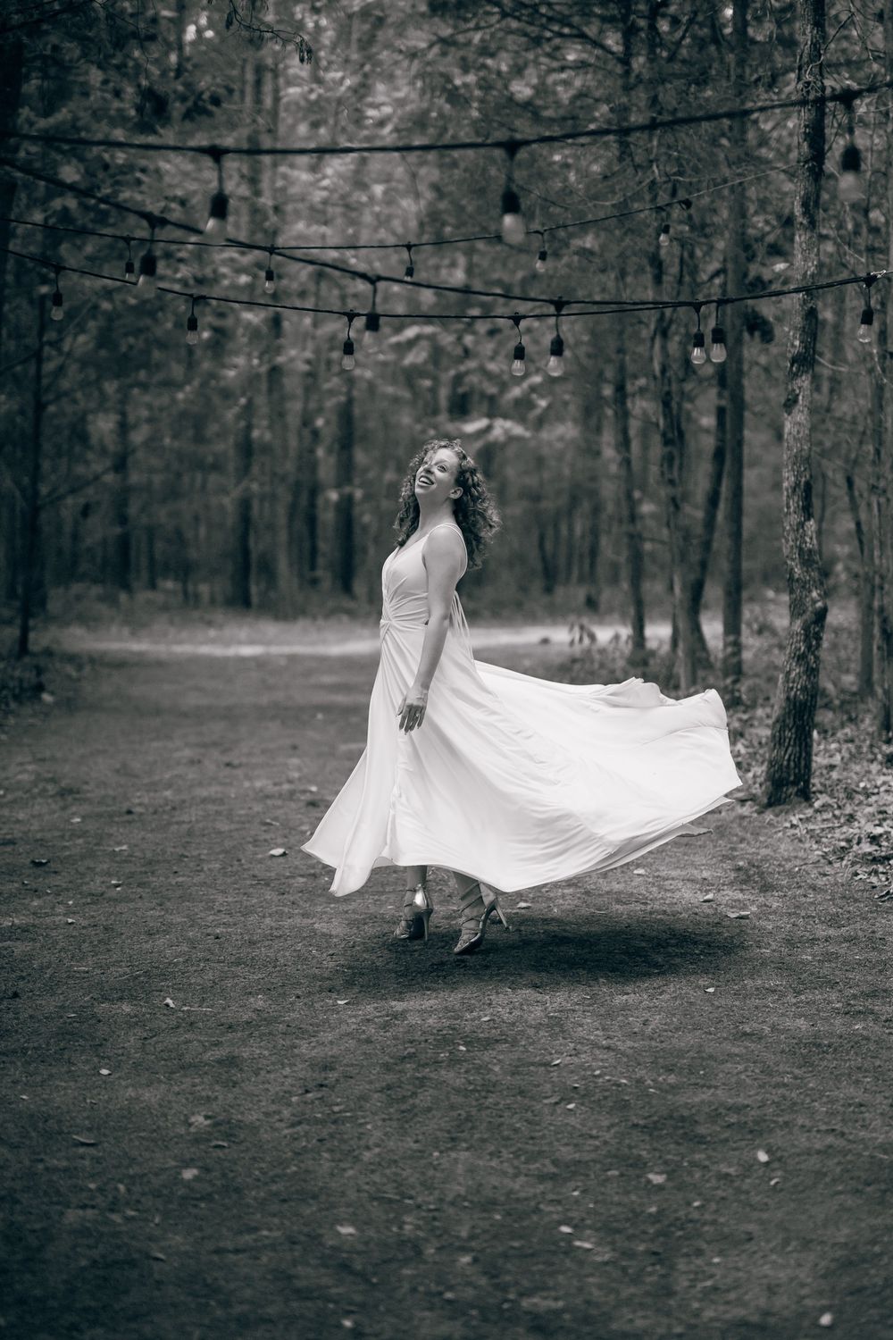 Black and white photo of a woman in a flowing white dress twirling in a forest path with string lights overhead.