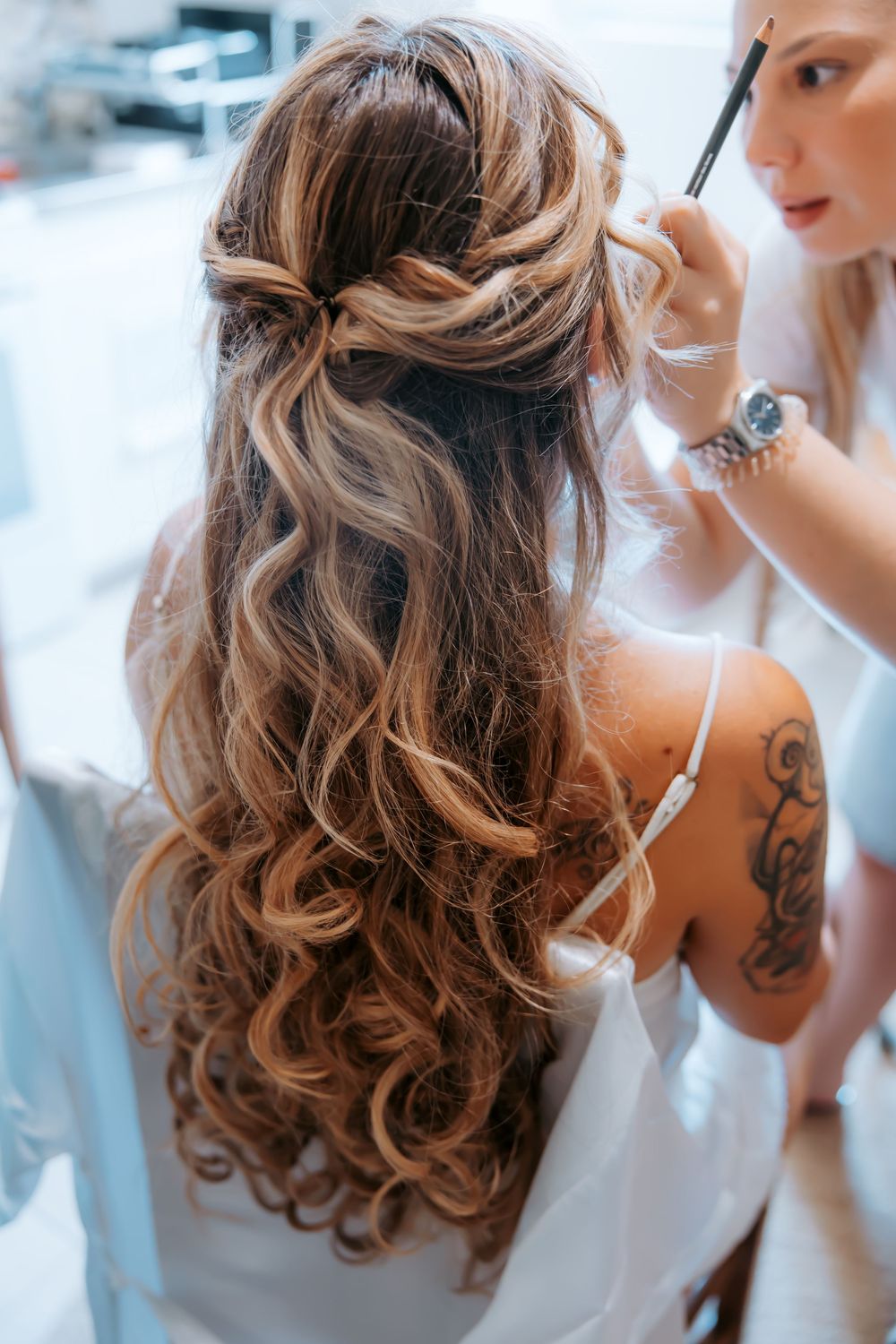 Bride with elegant half-up curly hairstyle being prepared by makeup artist with tattoo on arm.
