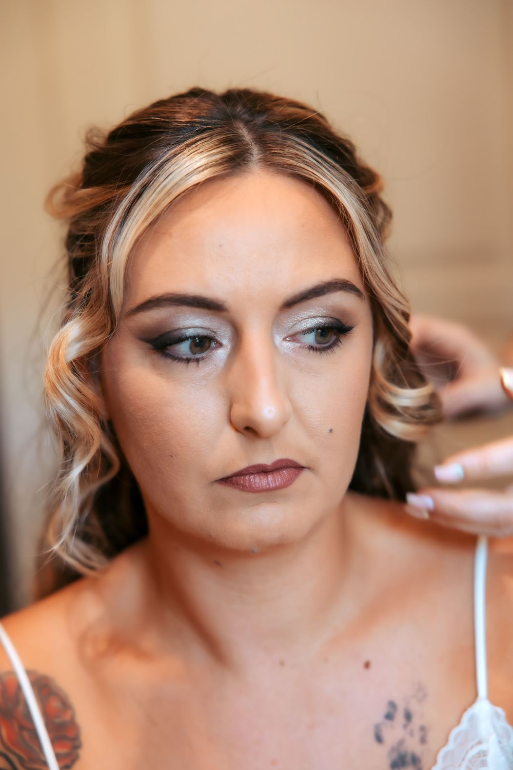 Bride getting makeup applied, featuring smoky eye shadow, dark lips, highlighted hair, and visible tattoo.