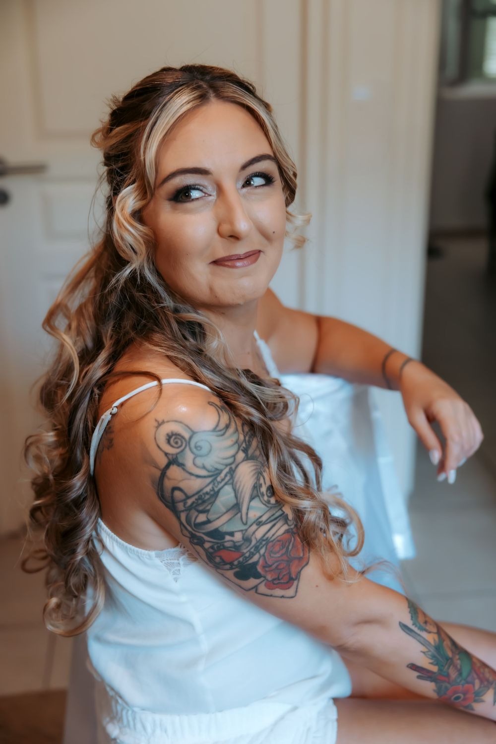 Young woman with curly blonde hair, blue eyes, and colorful tattoos on her arm, wearing a white top, smiling softly.