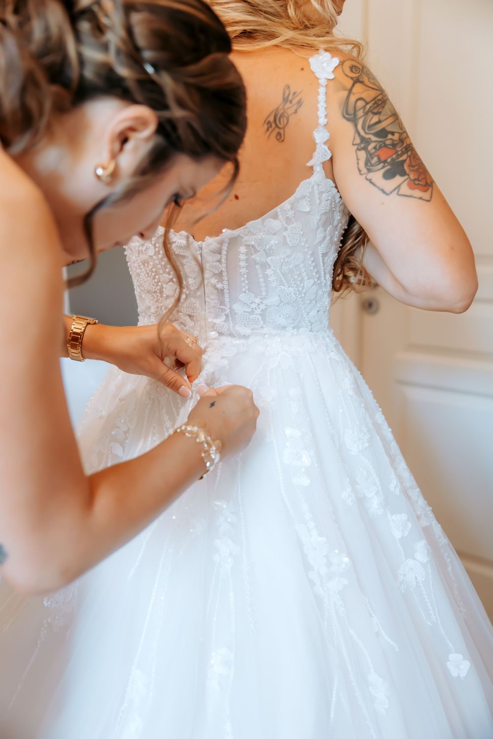 Bridesmaid helping bride button up white lace wedding dress from behind, showing tattoos and pearl bracelet.