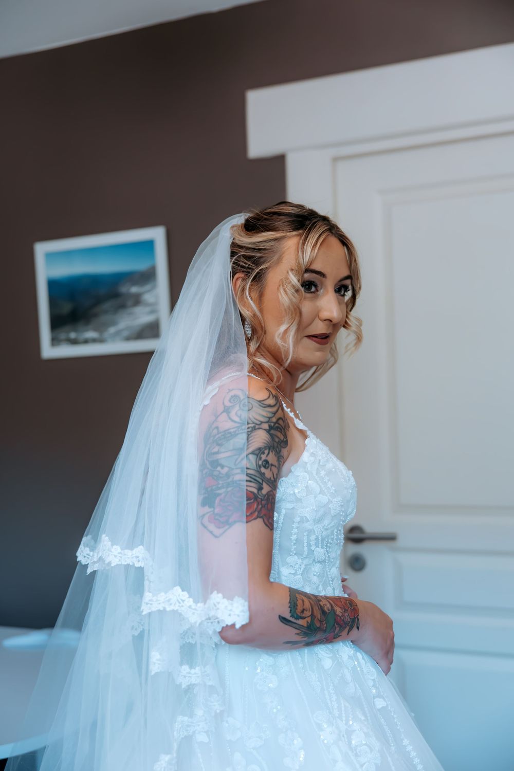 Bride in white lace wedding dress and veil with colorful arm tattoos poses elegantly indoors.