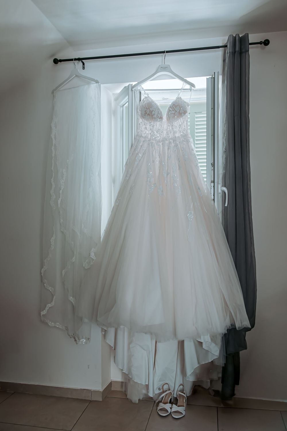 Elegant white lace ballgown wedding dress hanging by a window with a veil and bridal heels below.