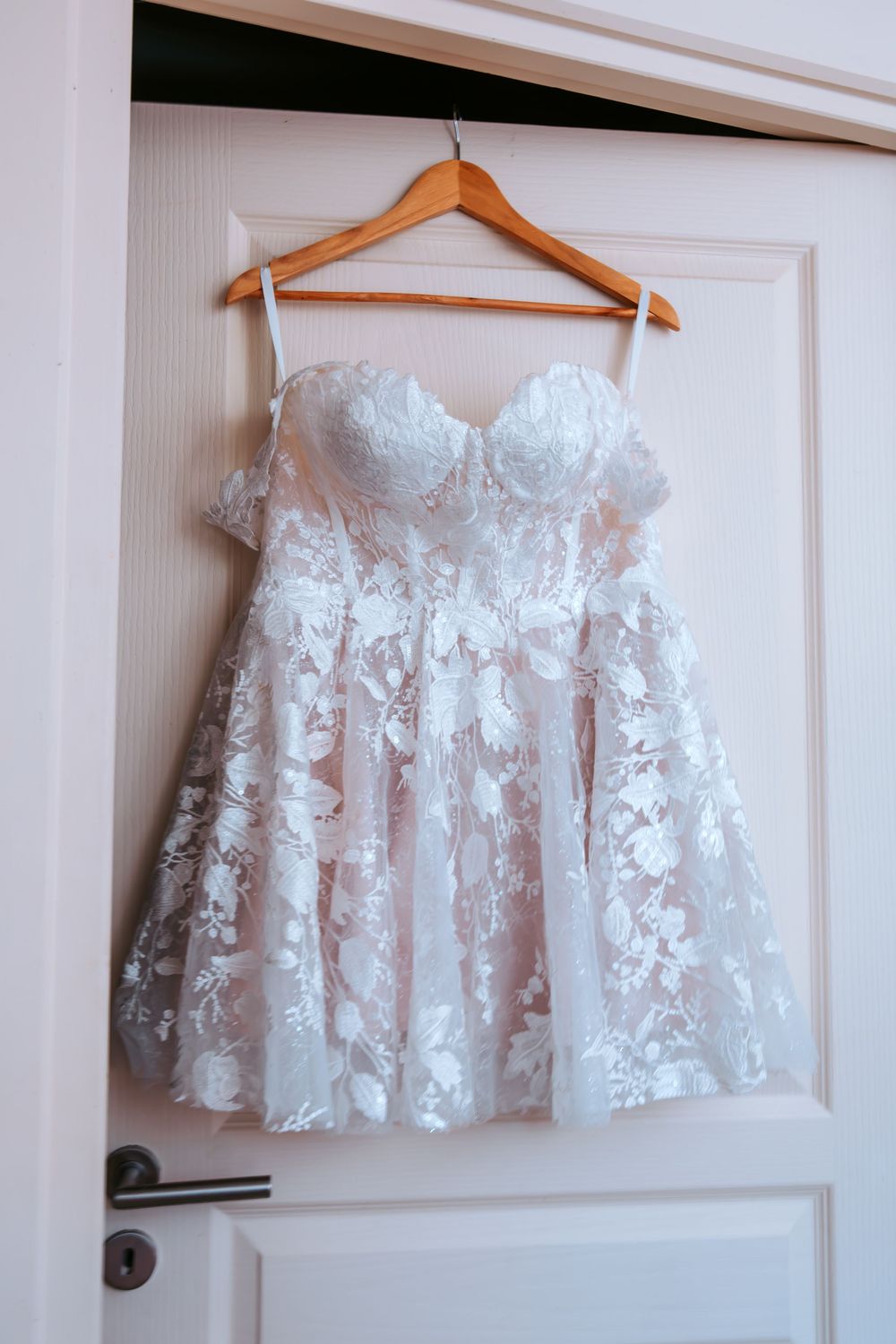 Short white lace wedding dress with sweetheart neckline hanging on wooden hanger on white door.