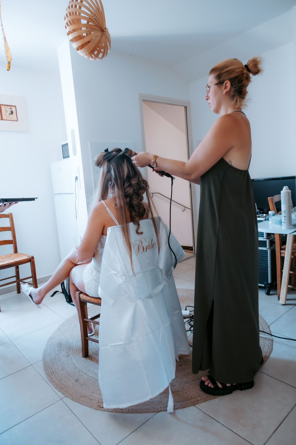 Hairstylist curling a bride's hair before wedding ceremony in a bright white room.