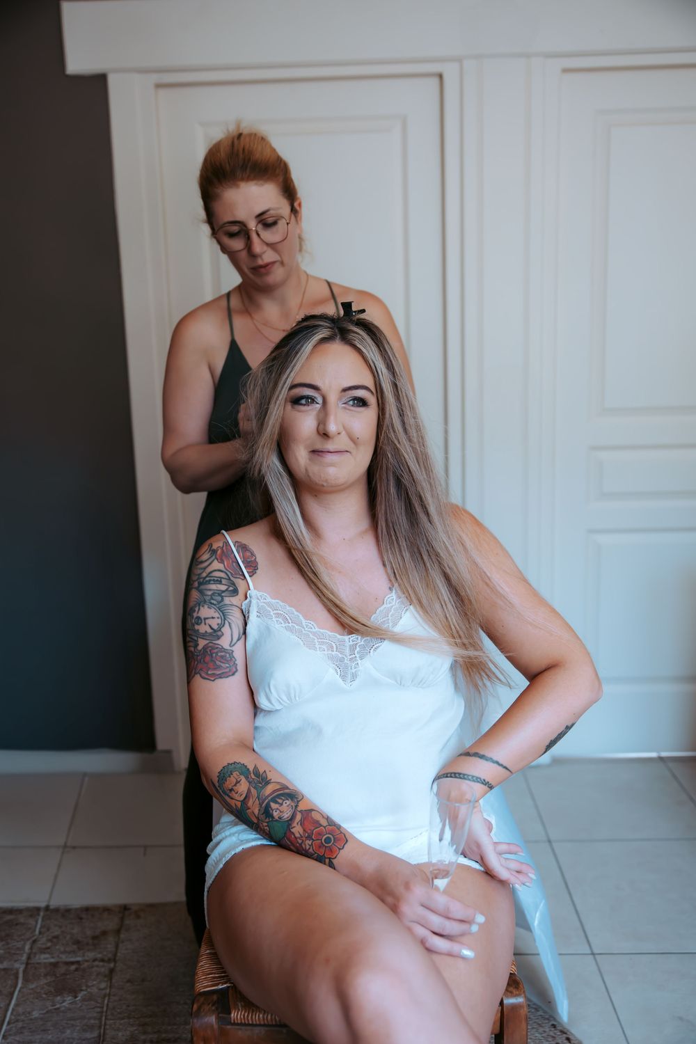 A tattooed bride in white sits while a hairstylist works on her long blonde hair indoors.