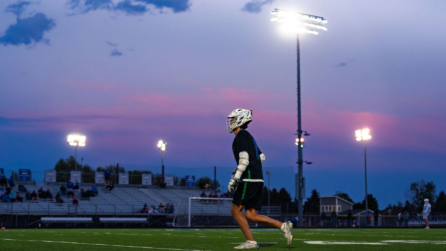Janesville lacrosse player walks across the field at dusk, lit by stadium lights under a purple and pink cloud-streaked sky.
