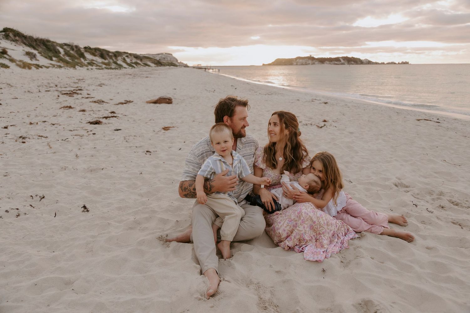 Family of four sitting on sandy beach at sunset, parents smiling with toddler and young girl in pink dress.