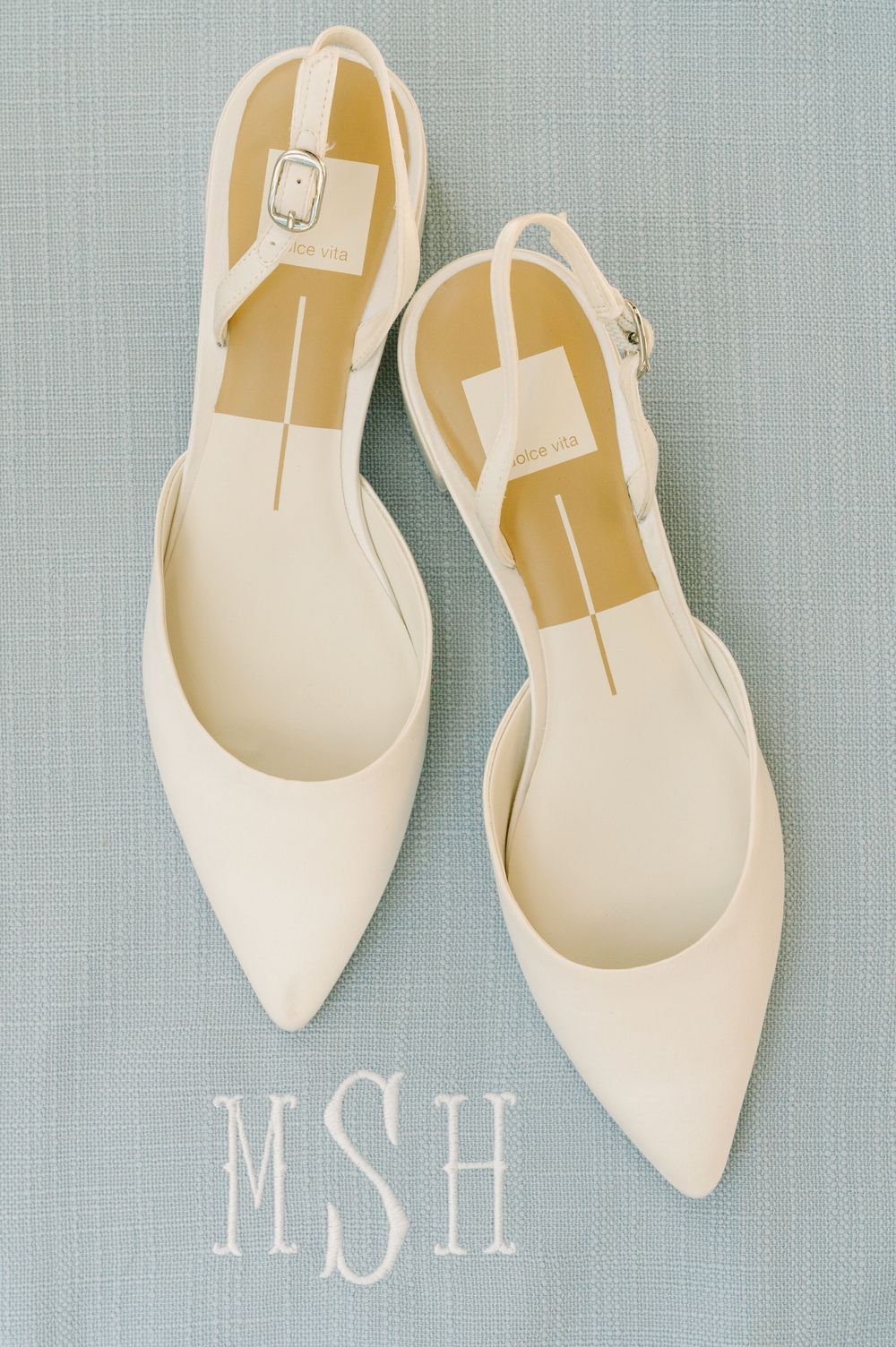 White pointed-toe slingback flat wedding shoes displayed on blue linen with MSH monogram.