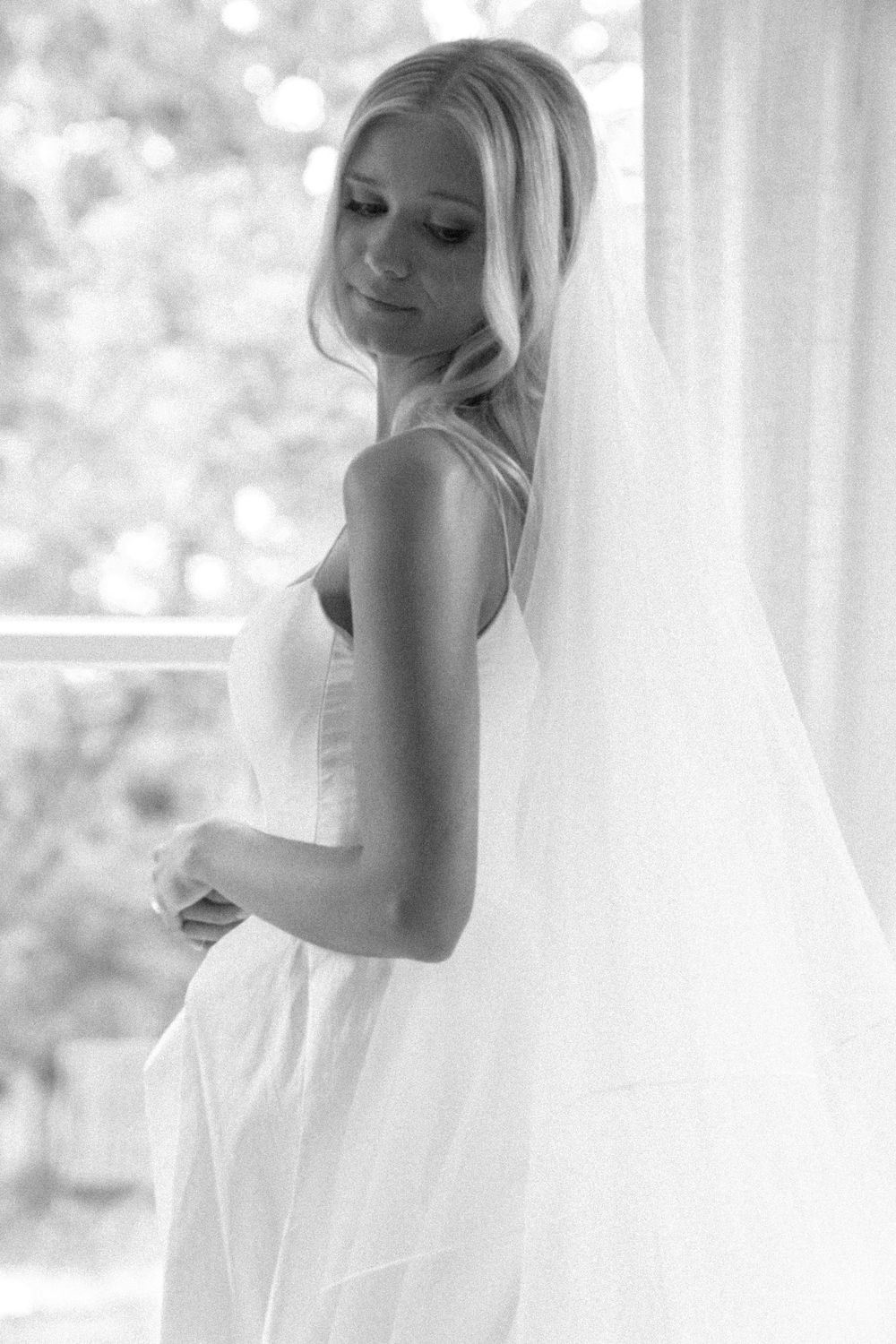 Elegant bride in white gown and veil posing by window in black and white wedding portrait.