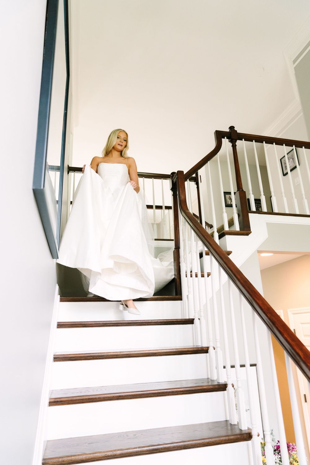 Bride in white gown descending elegant wooden staircase on wedding day.