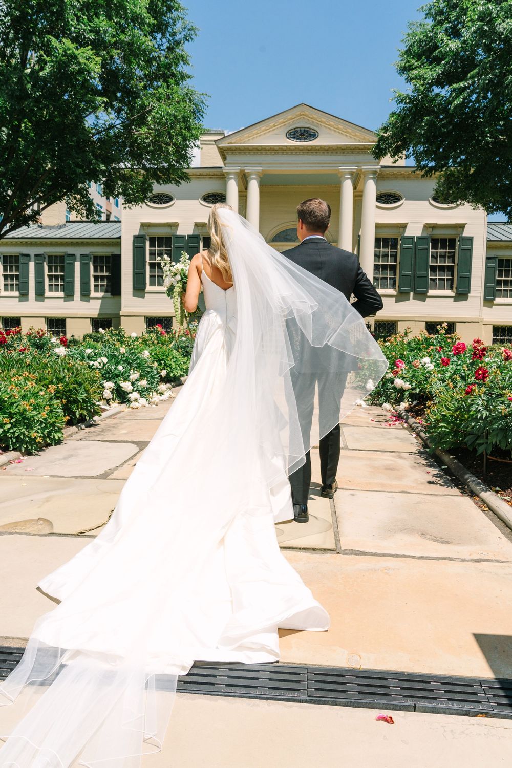 Bride and groom walking hand in hand toward a grand estate surrounded by lush gardens on their wedding day.
