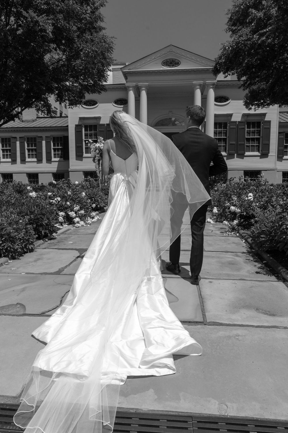 Black and white photo of a bride and groom walking toward an elegant estate, her veil flowing in the breeze.
