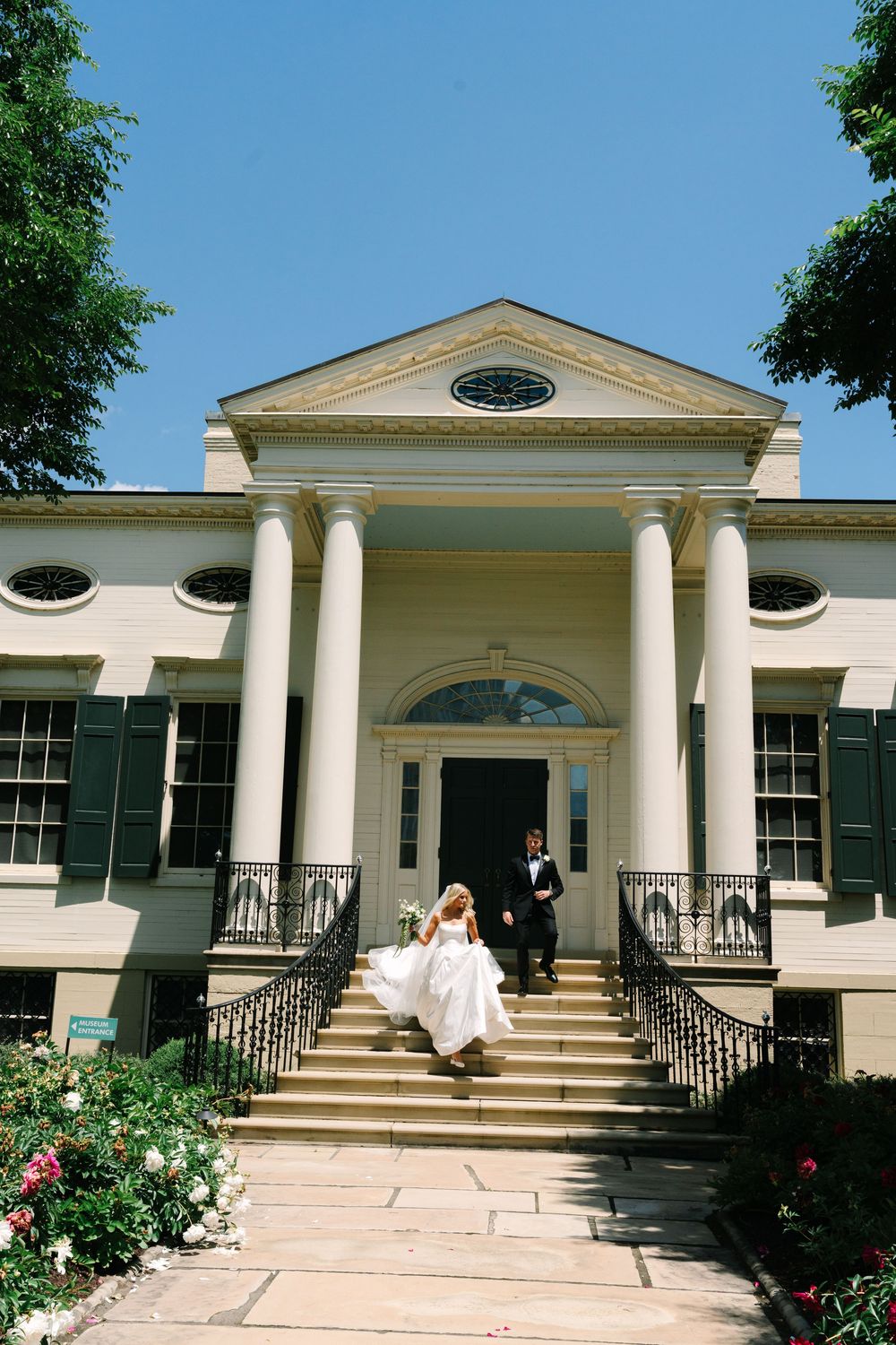 Bride and groom descend grand colonial mansion steps in wedding attire under blue sky.
