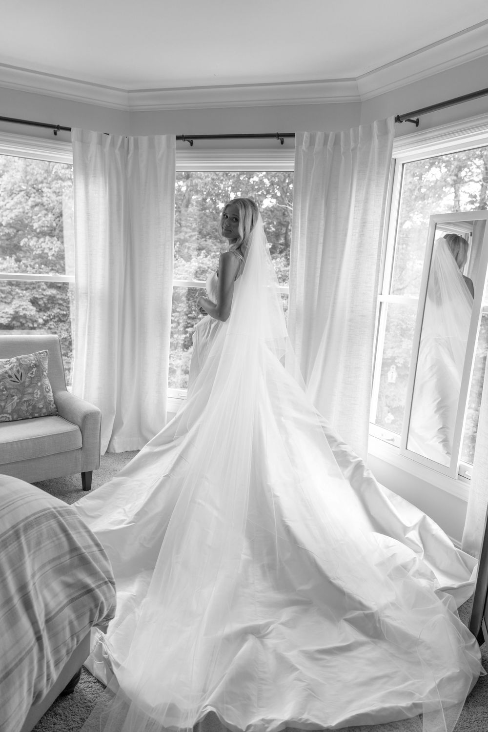 Bride in elegant white gown with long train stands by bay window in black and white wedding photo.