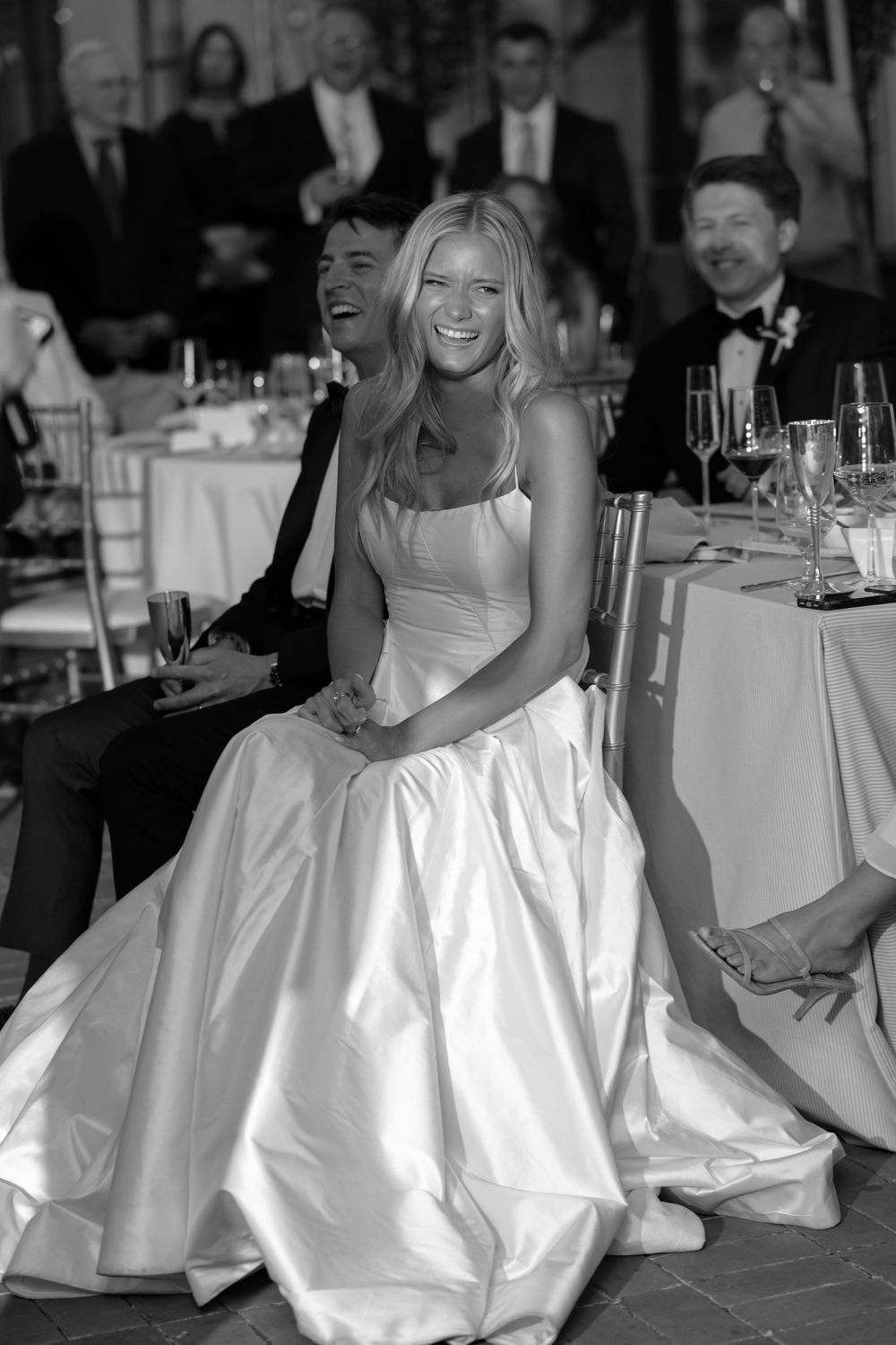 Bride laughing joyfully in strapless ball gown at wedding reception surrounded by guests.