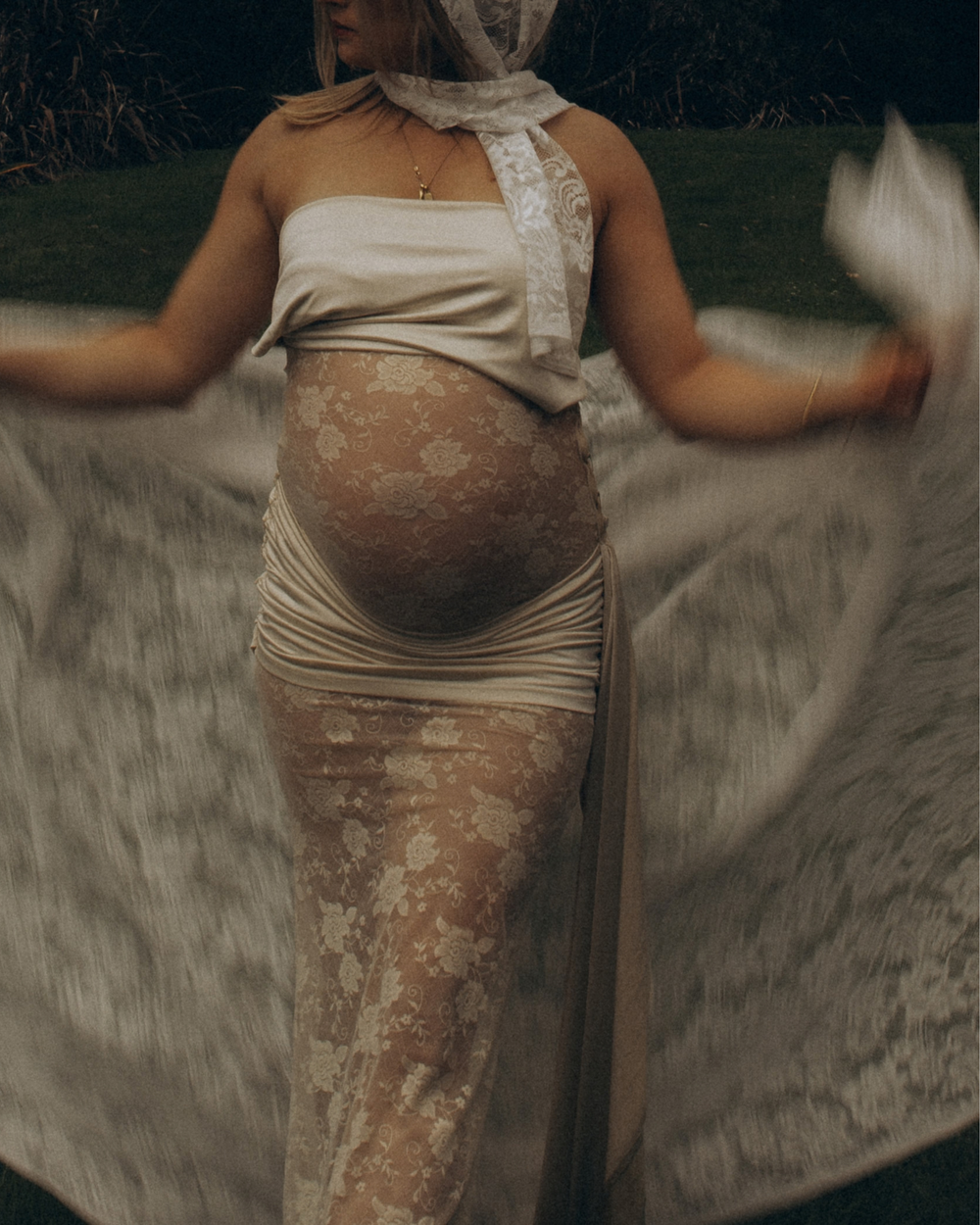 Pregnant woman in sheer lace maternity gown holding flowing fabric outdoors during elegant maternity photoshoot.