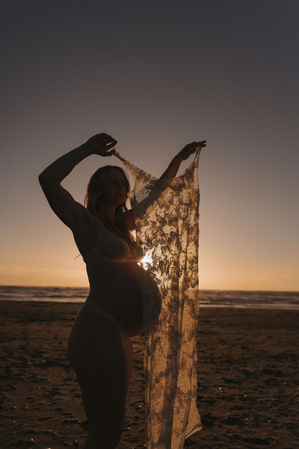 Pregnant woman holding lace fabric at sunset on beach, silhouetted against golden ocean horizon.