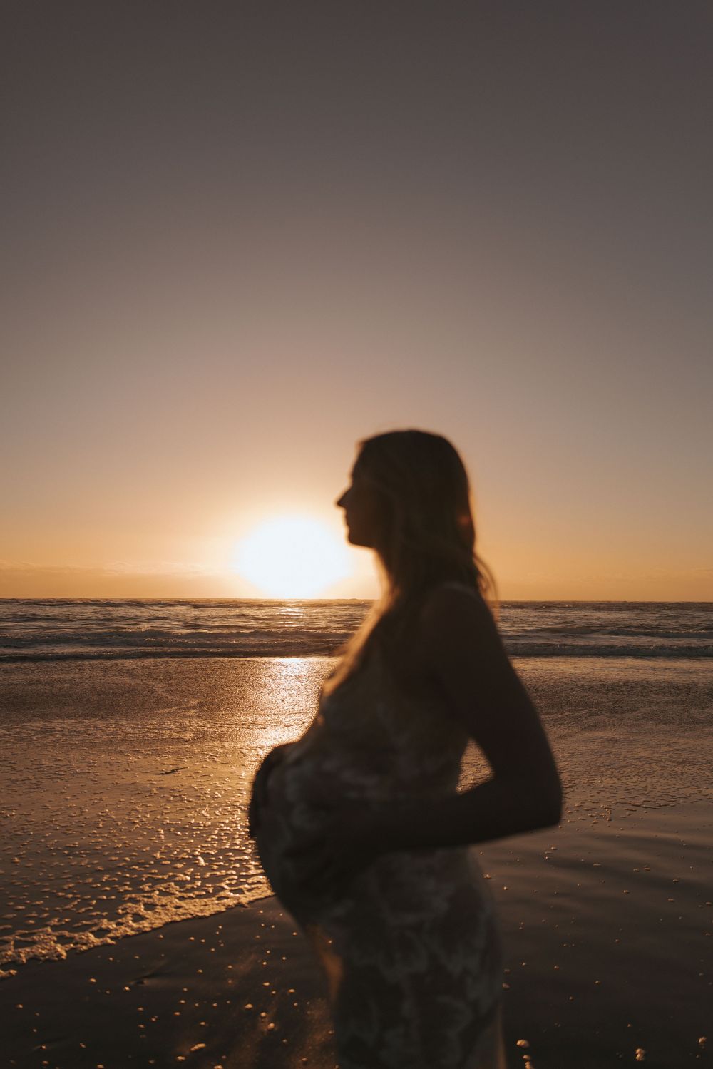 Silhouette of a pregnant woman standing on a beach at golden sunset, waves reflecting warm light behind her.
