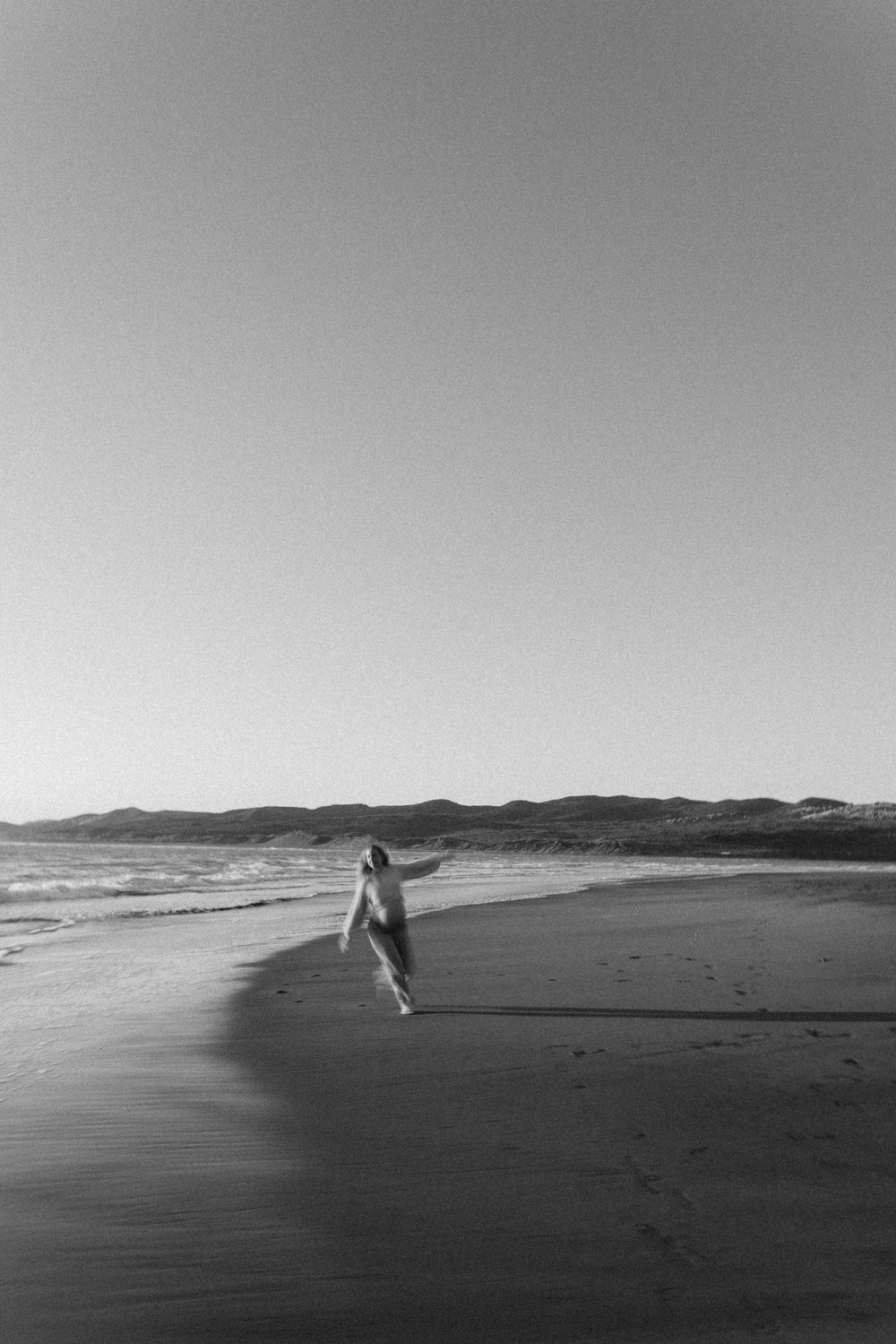 Black and white photo of a person walking along a serene beach with ocean waves and distant mountains.