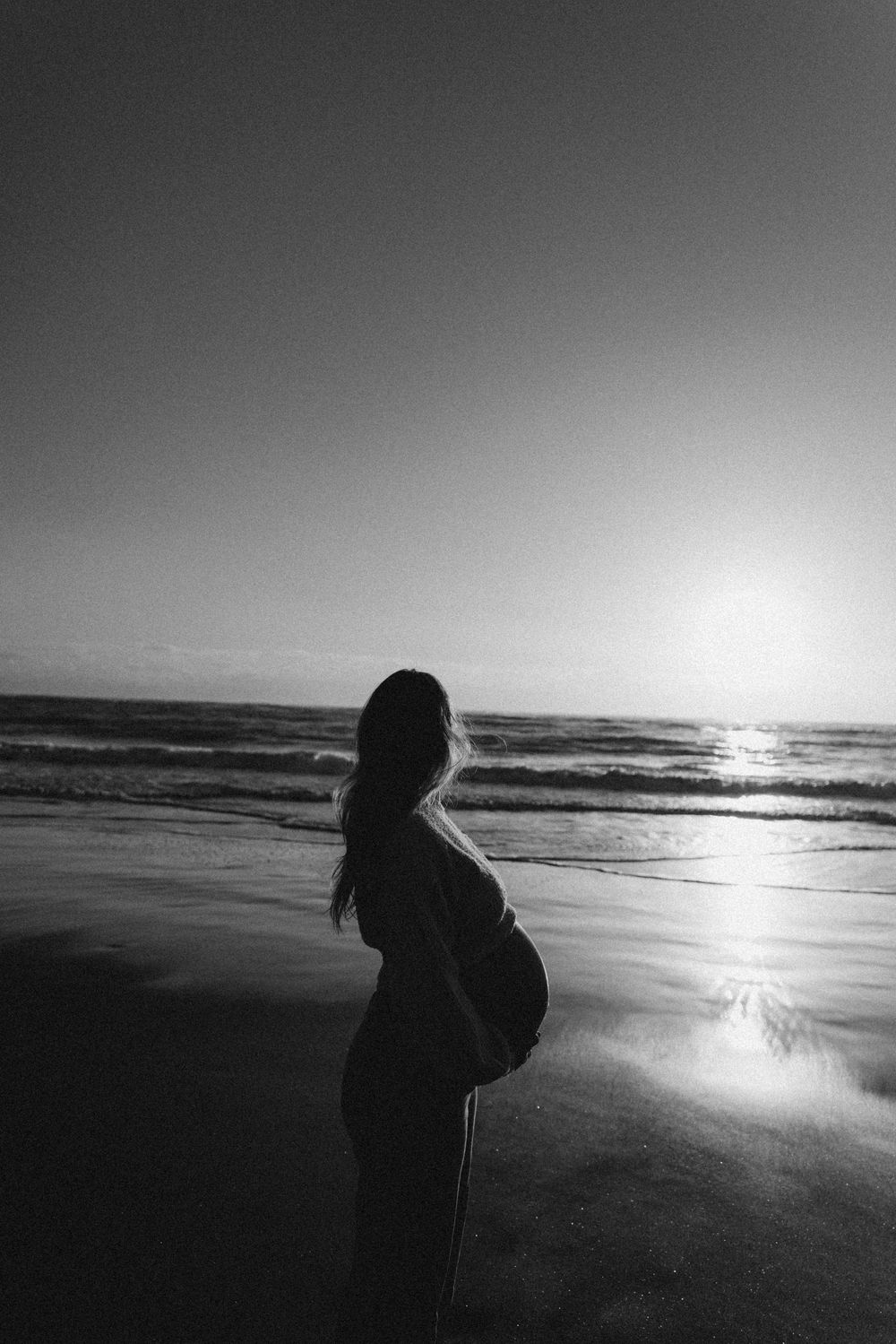 Black and white photo of a pregnant woman silhouetted against a serene ocean sunset on the beach.