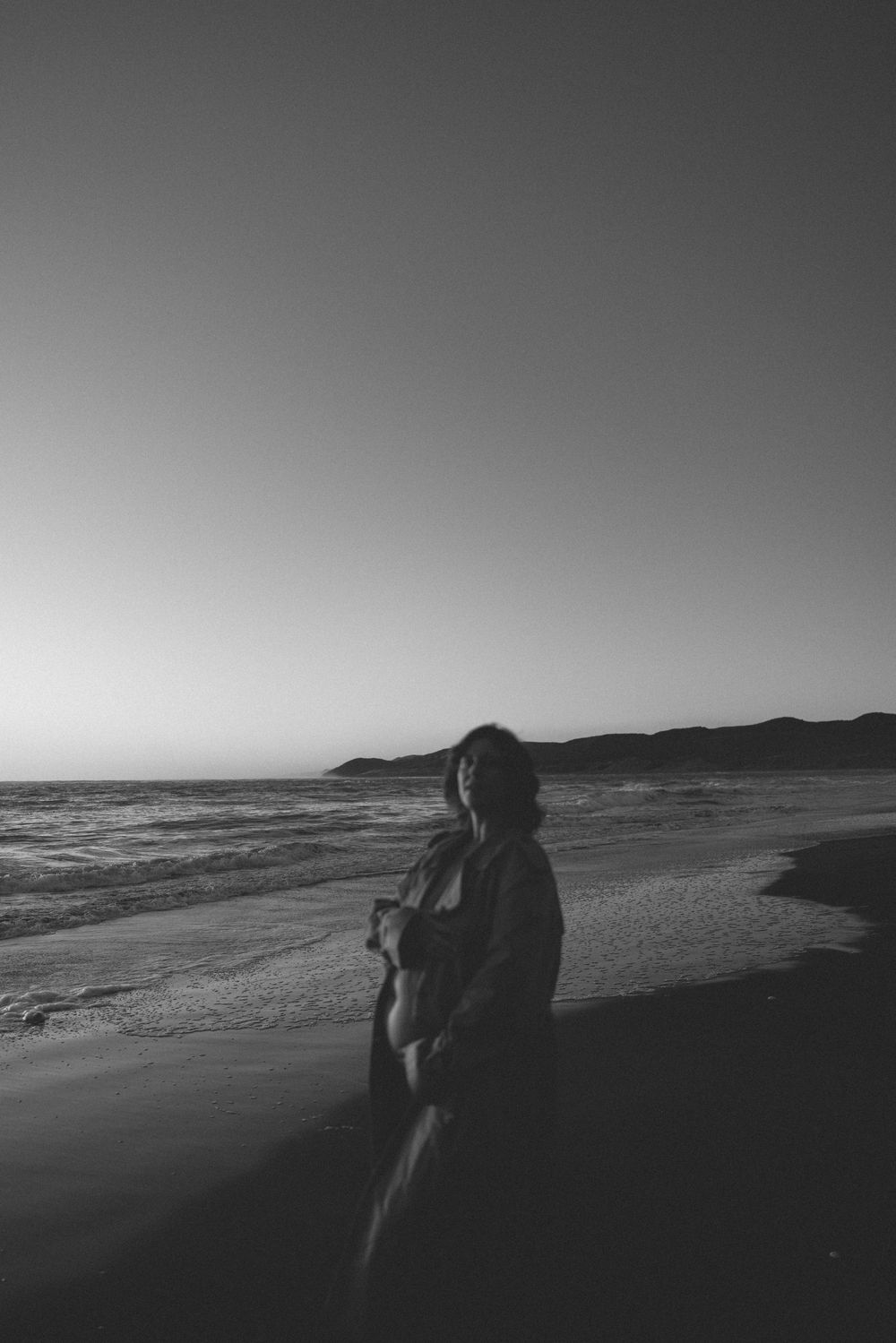 Black and white photo of a person standing on a beach at dusk with ocean waves and mountains in the background.