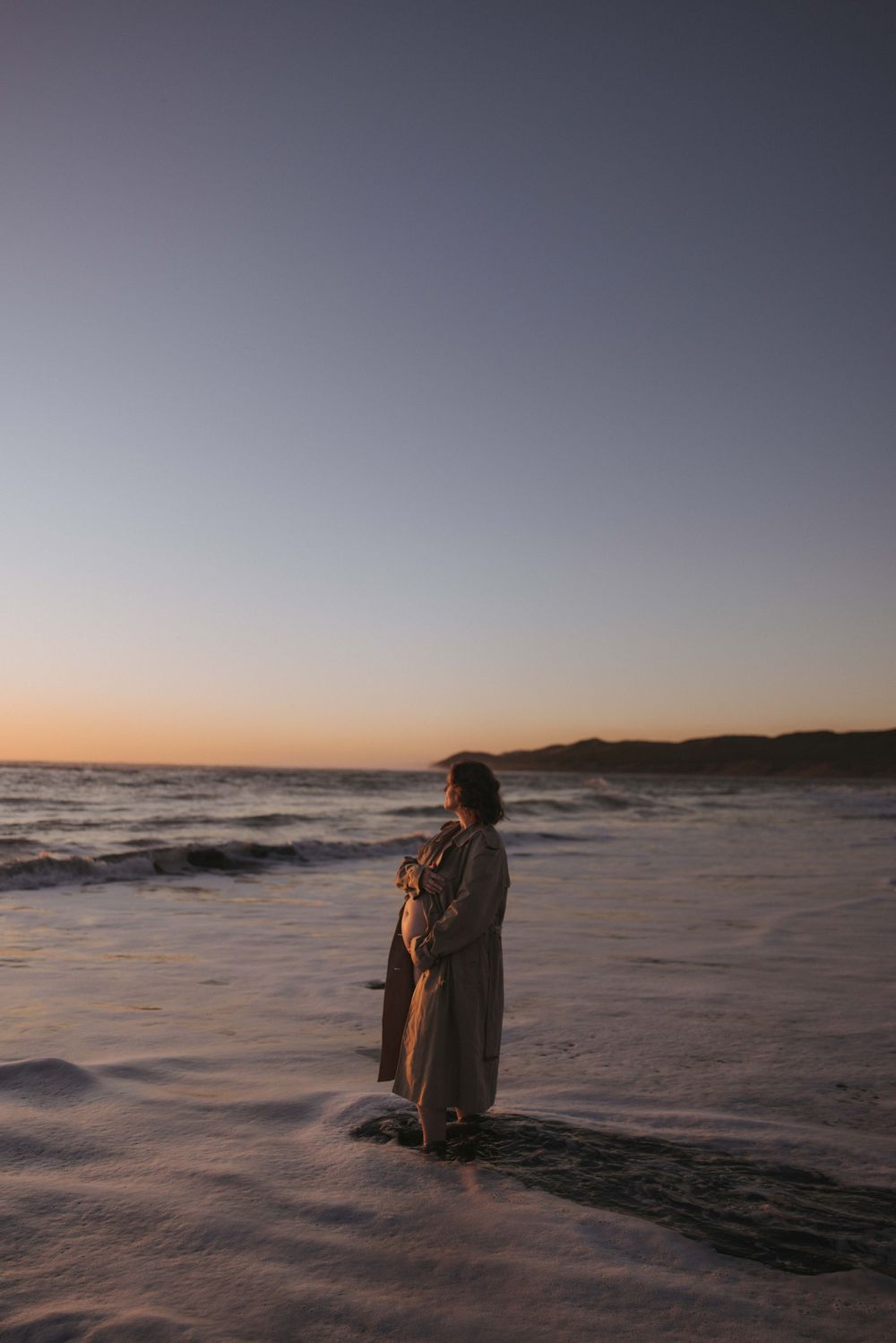 Woman in long coat standing on beach at sunset with ocean waves and hills in background.