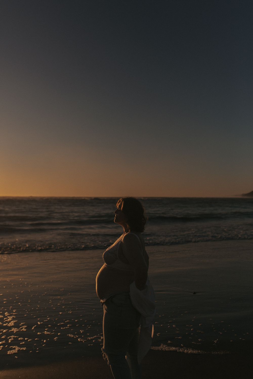 Pregnant woman silhouette standing on beach at golden sunset, cradling baby bump while ocean waves wash ashore.