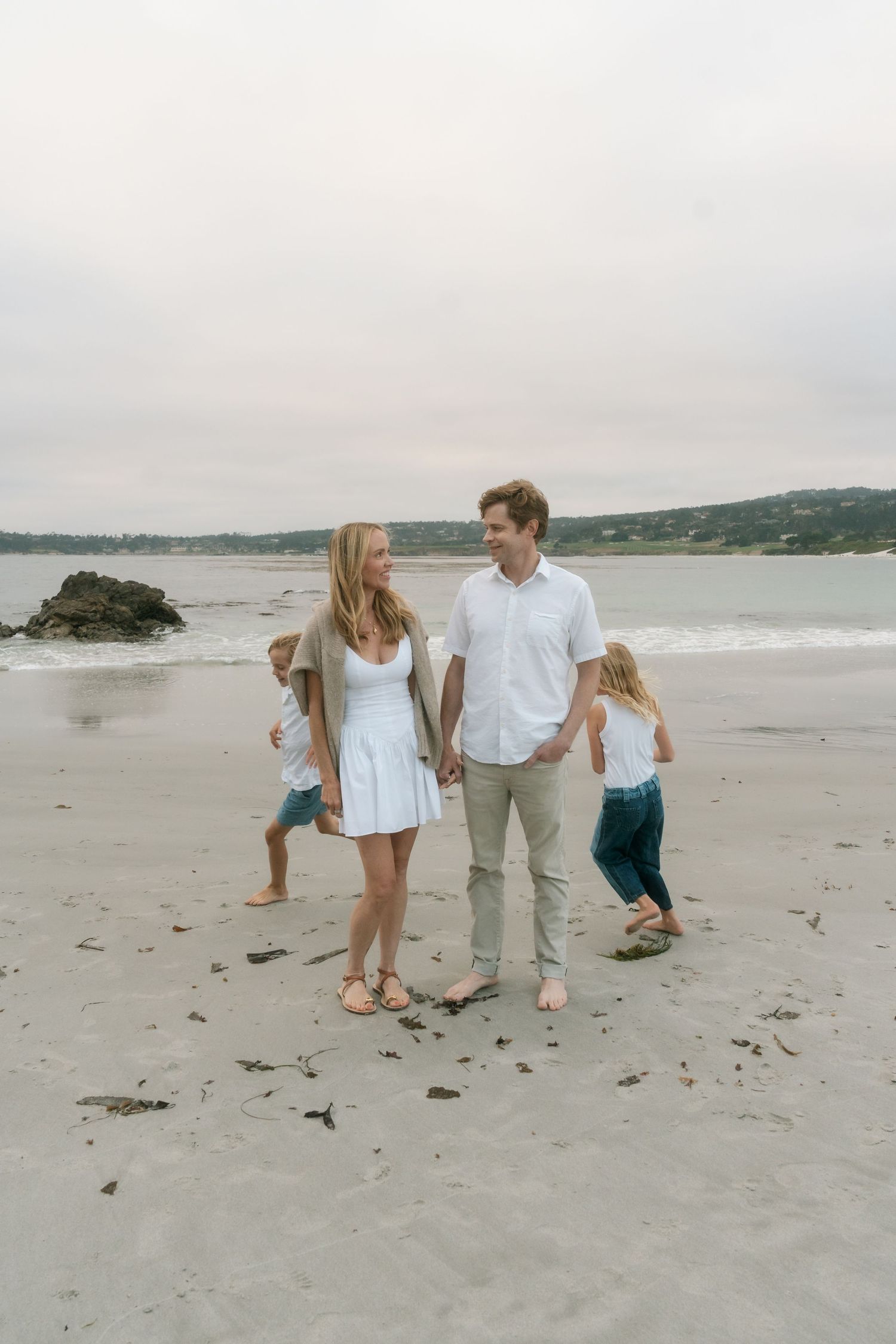 A happy family of four walks hand-in-hand on a sandy beach, with two young children playing around them.