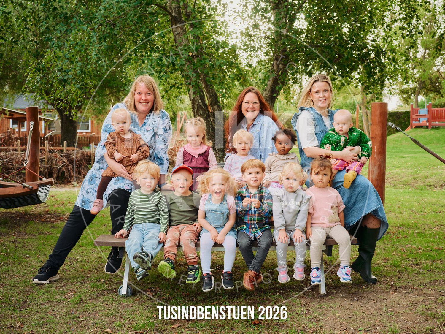 Three women pose outdoors with a large group of toddlers on a bench, text reads Tusindbenstuen 2026.