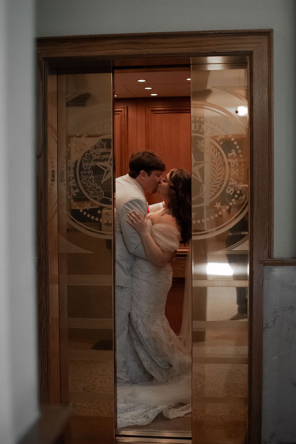 Newlyweds kiss inside an elevator with decorative glass doors during their Fort Worth courthouse wedding portraits.