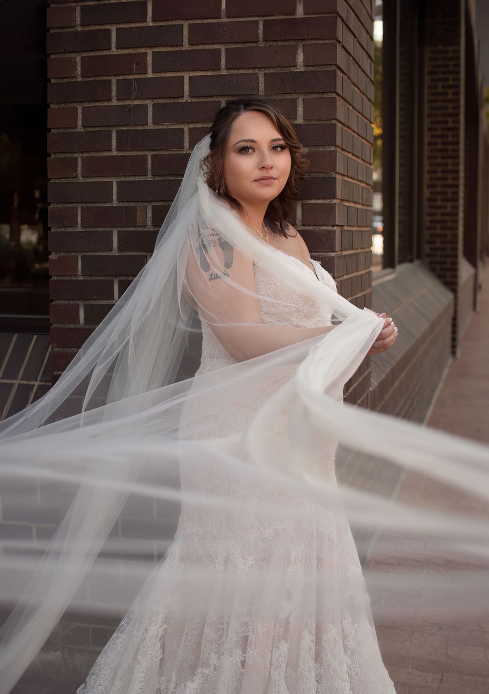 Bride in a lace wedding dress and flowing veil poses beside a brick building during her downtown Fort Worth courthouse portraits.