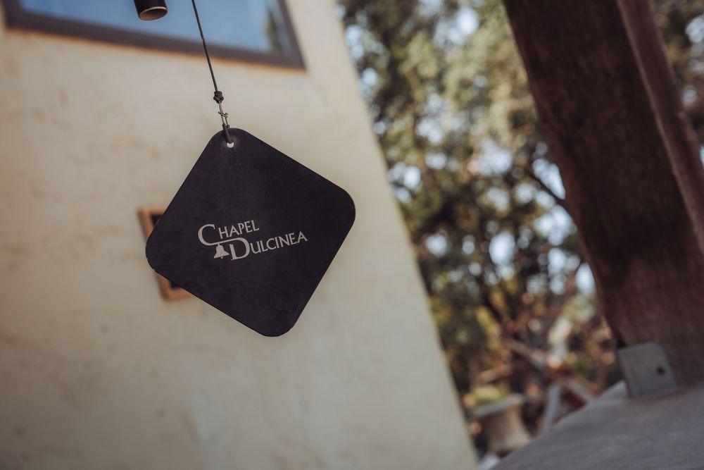 A hanging black sign reading Chapel Dulcinea against a blurred outdoor background with trees and a building.