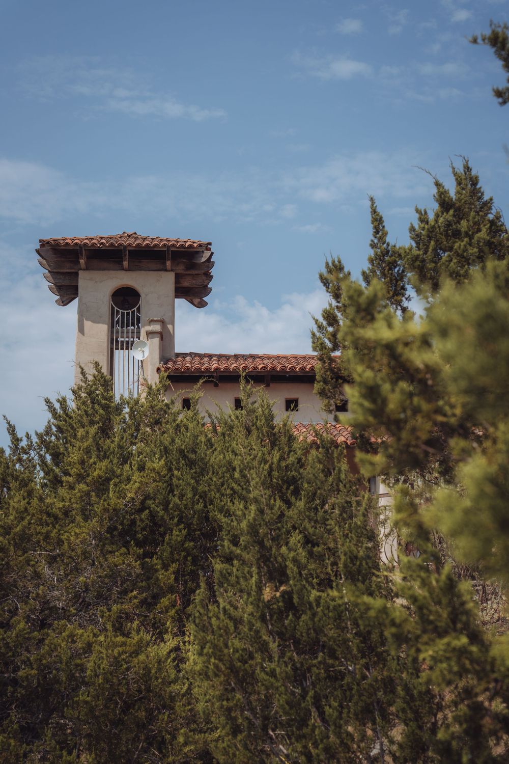 A Spanish Colonial Revival tower with red tile roof rises above lush green cypress trees against a blue sky.