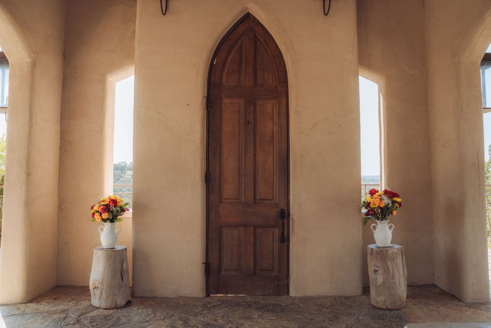 Rustic wooden arched door flanked by floral arrangements on tree stump pedestals at a beige stucco chapel entrance.