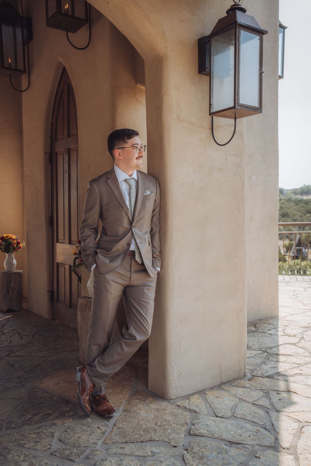 Elegant groom in tan suit leaning against stone pillar on rustic venue porch with vintage lantern.