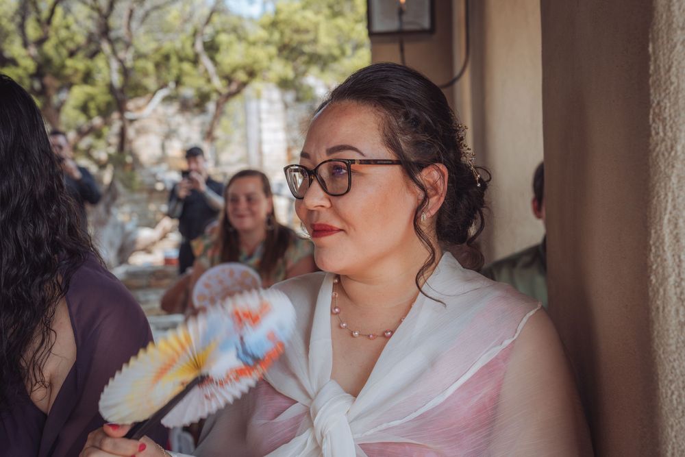 Woman with glasses holding a decorative fan at an outdoor event, wearing a pink outfit on a sunny day.