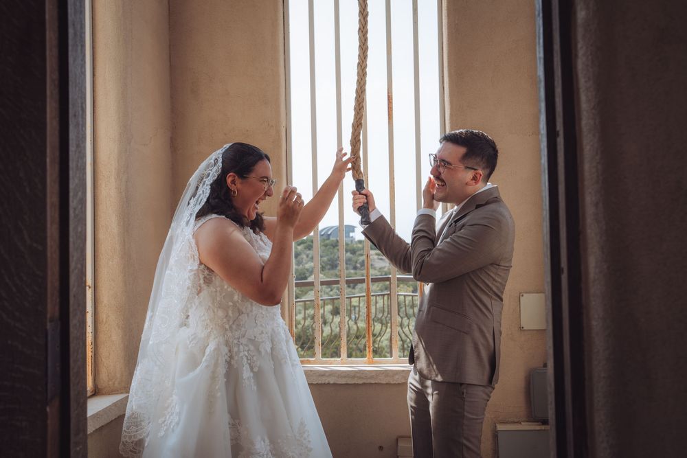 Bride and groom holding a rope together near a sunlit window in a rustic venue on their wedding day.