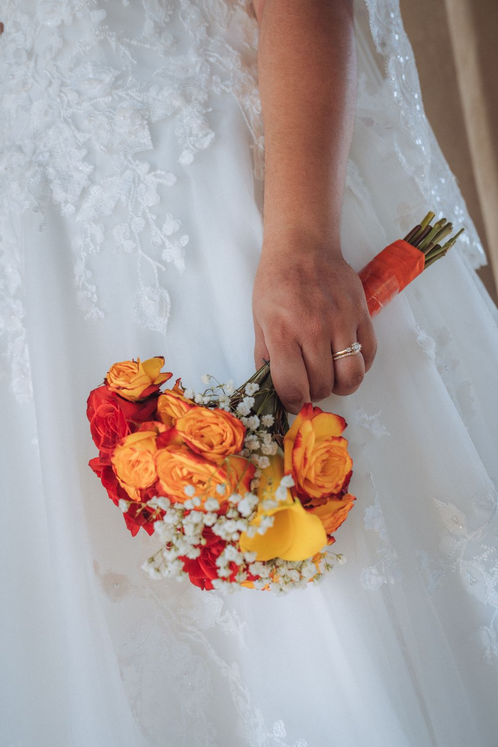 Bride holding a vibrant wedding bouquet of orange roses, red flowers, and white baby's breath against a white lace gown.