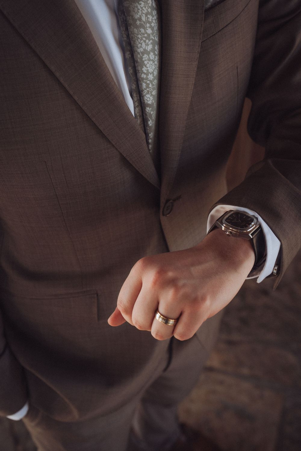 Man in brown suit with gold wedding band and luxury wristwatch, elegant formal attire close-up.