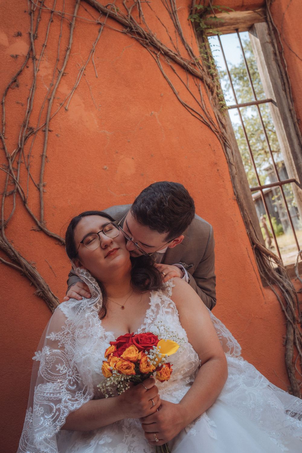 Groom kissing bride on cheek near orange wall with vines, bride holding colorful rose bouquet in lace wedding dress.