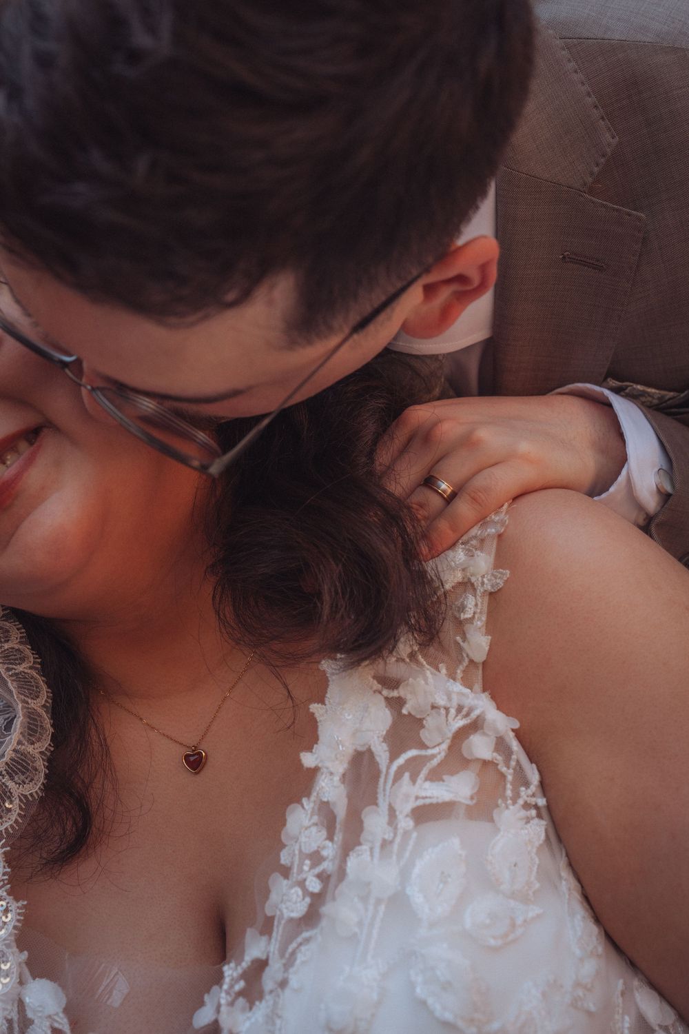 Groom in glasses kissing bride on cheek, wearing lace wedding dress and heart necklace, with wedding ring visible.