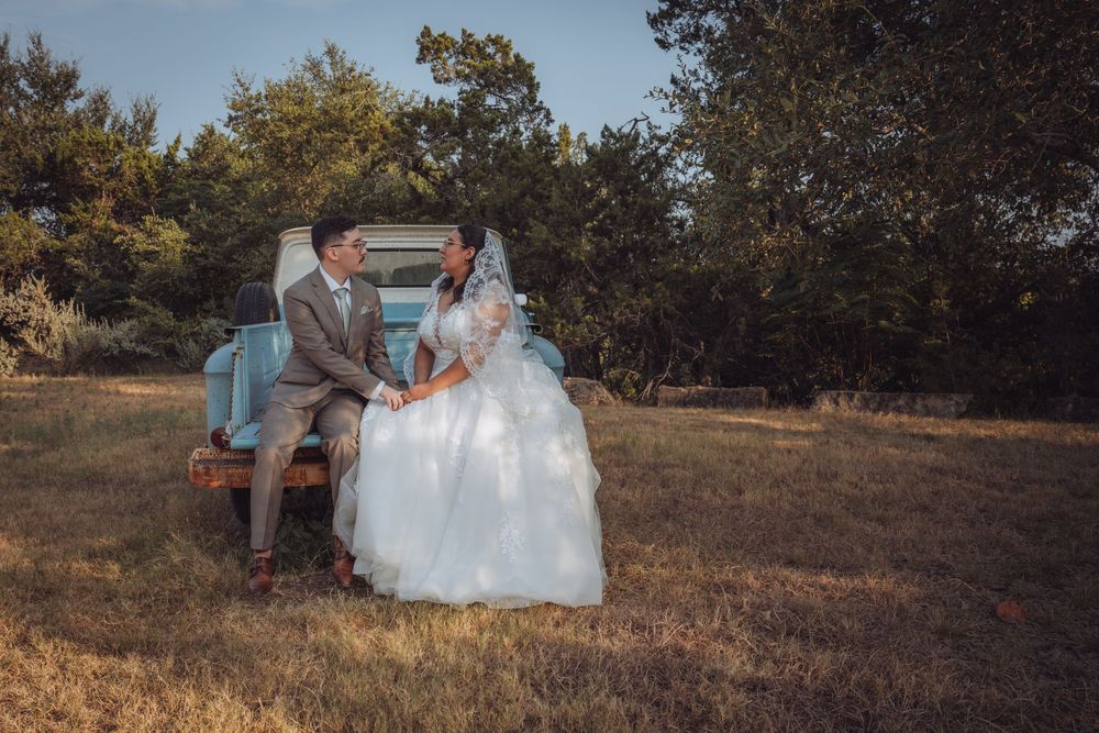 A bride and groom sit together on a vintage blue truck in a rustic outdoor field surrounded by trees on their wedding day.