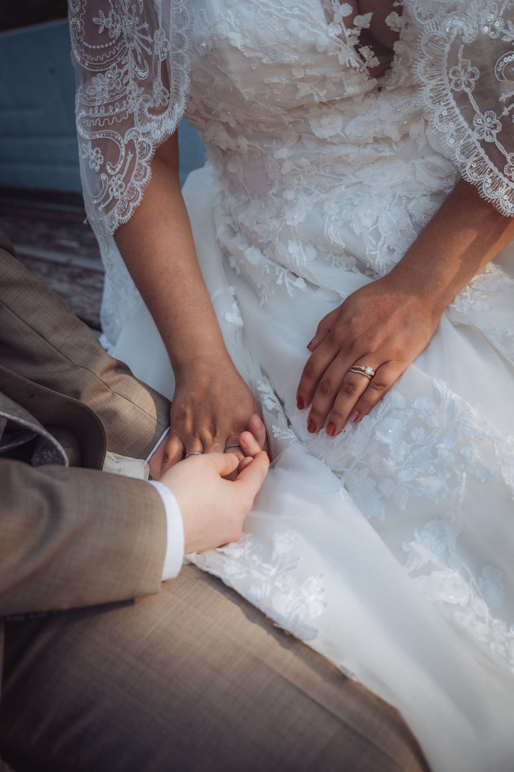 Bride and groom holding hands, bride wearing lace wedding dress and gold ring, groom in gray suit.