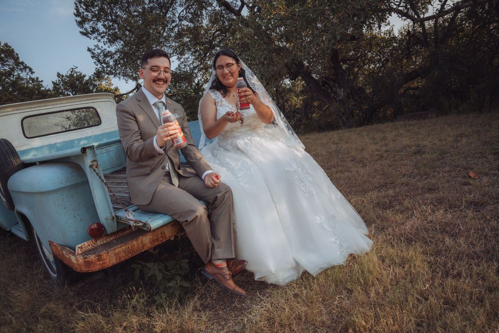 Bride and groom sitting on vintage blue truck tailgate, laughing and holding drinks outdoors.